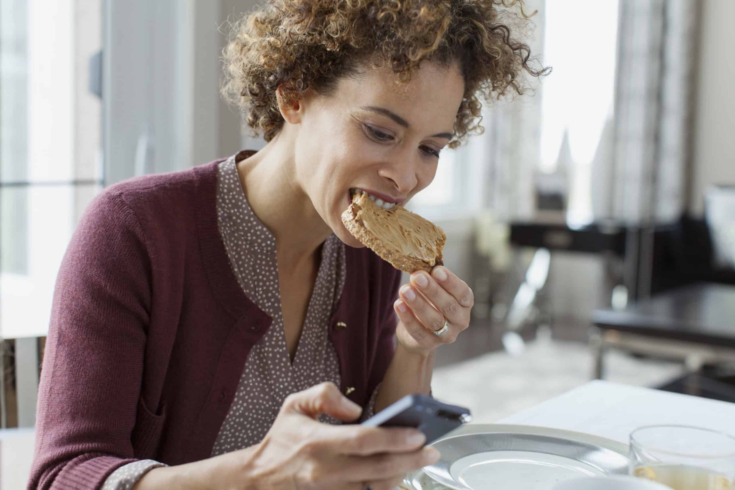 Woman eating toast while checking smartphone.