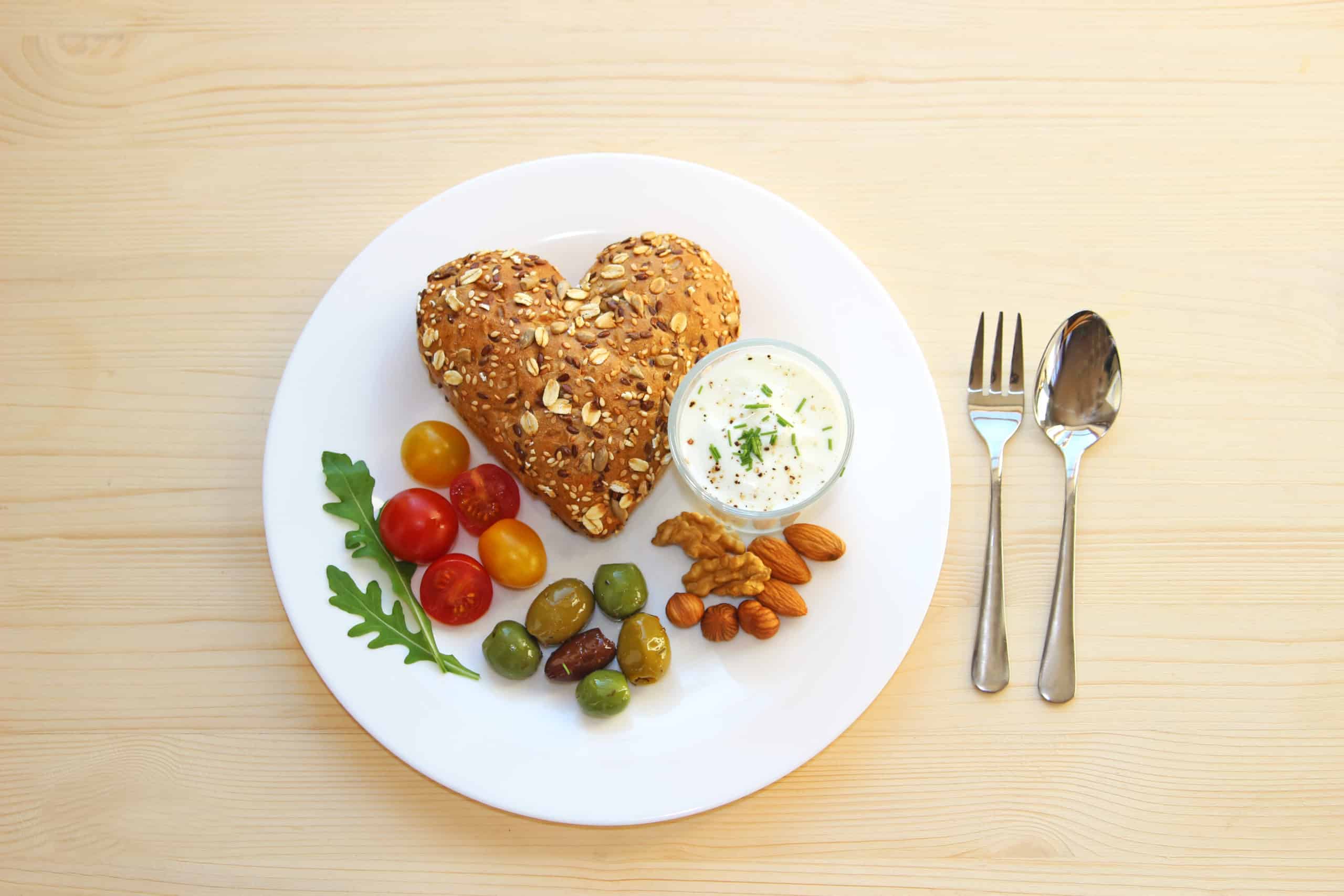 healthy food for your heart, plate with wholegrain bun in heart shape, yogurt, olives, nuts and tomatos on bright wood table
