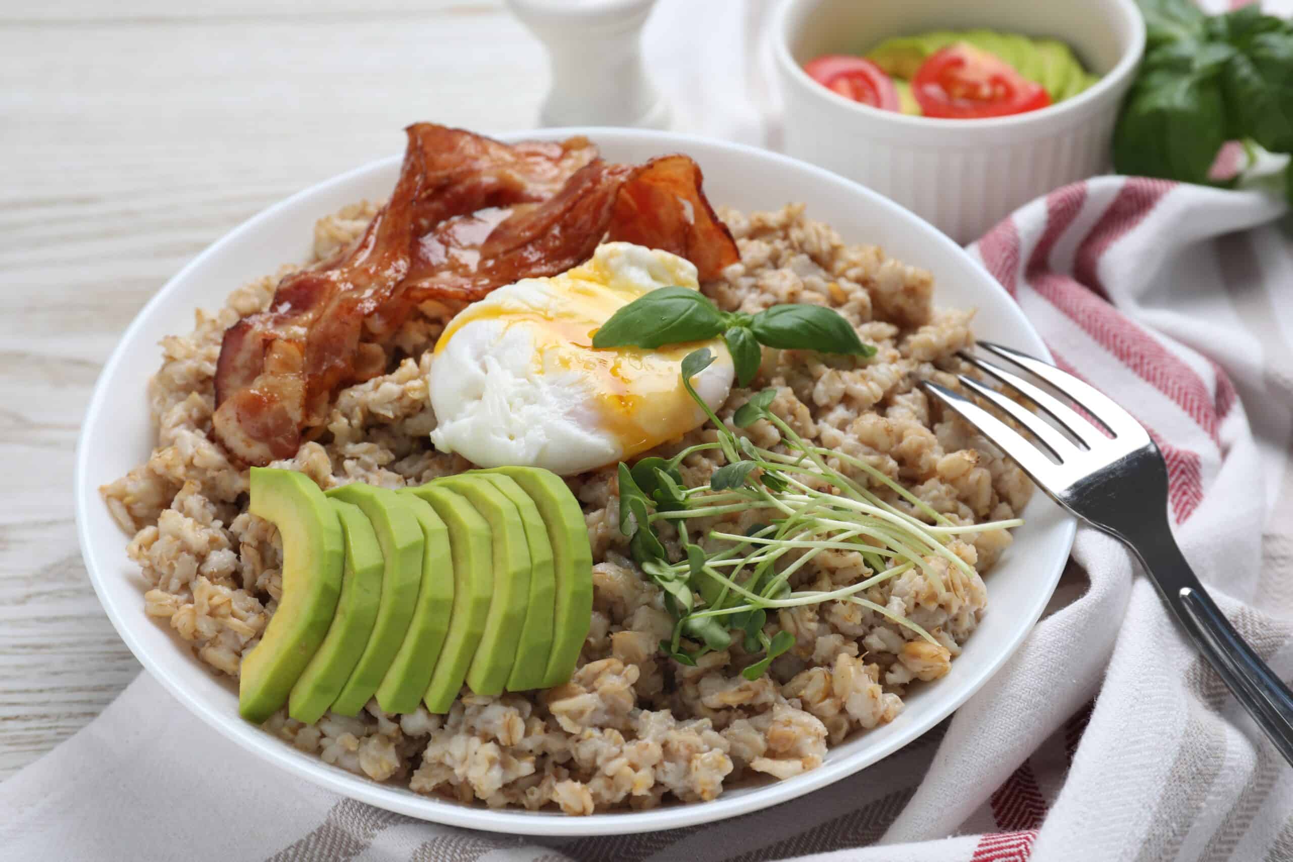 Delicious boiled oatmeal with poached egg, bacon, avocado and microgreens on table, closeup